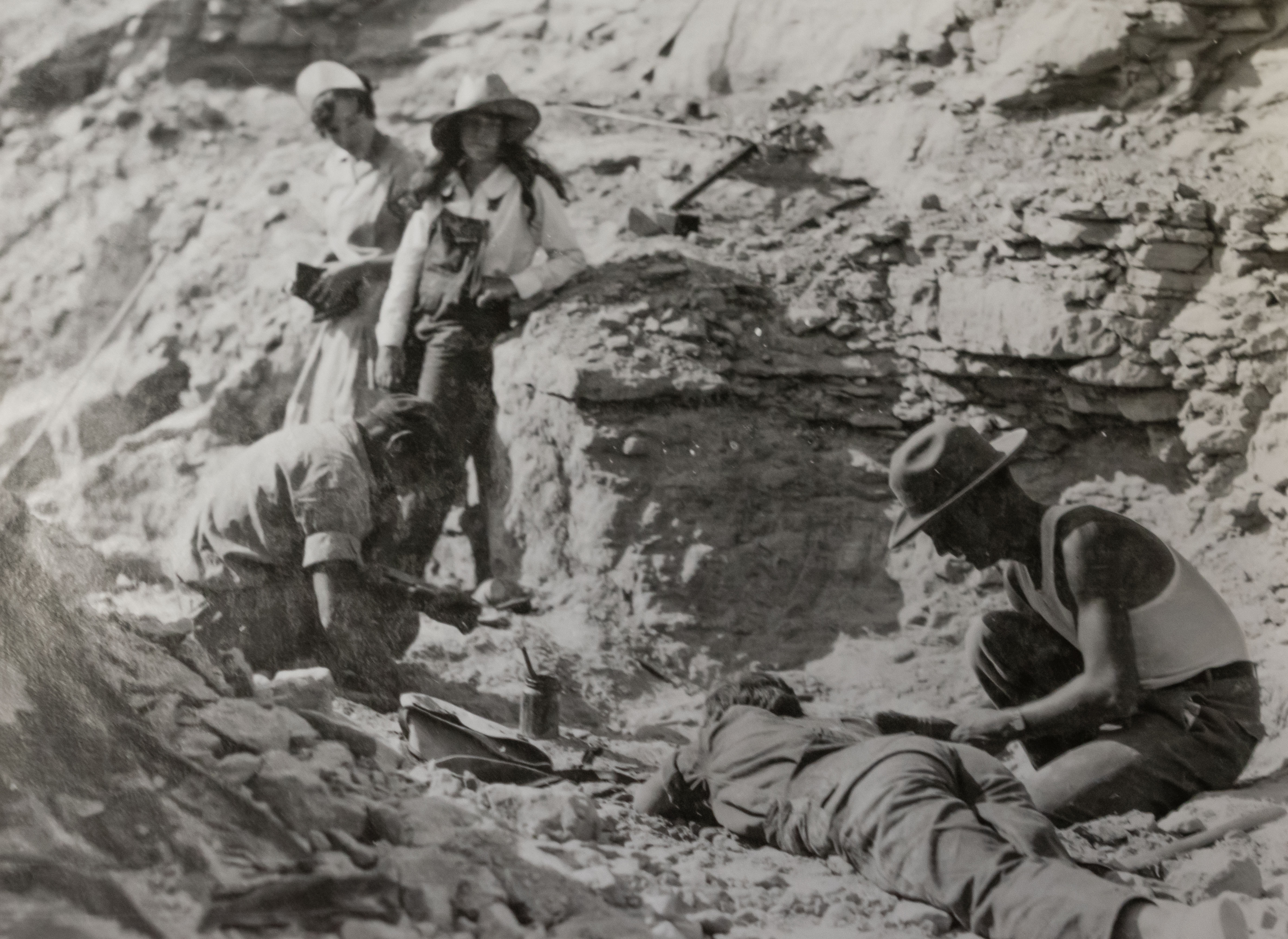 BW: 2 young women lean against a boulder in the hills while another girl lies on stomach in dirt looking in a hole. 2 young men with tools sit near the woman.