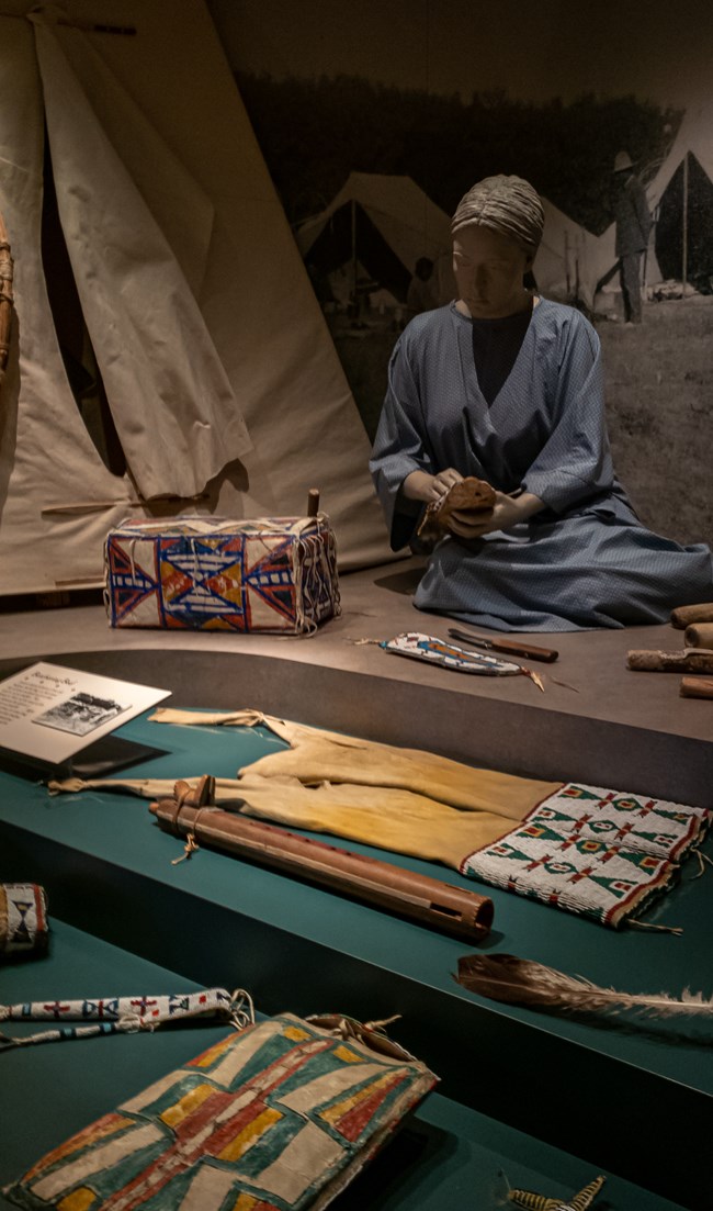A mannequin of a woman in a blue dress works next to a tipi. Native American artifacts made of leather, wood, and colorful beads surround her in the foreground.