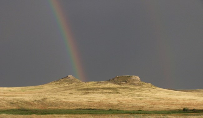 A pointed hill on the left and a flat-topped hill on the right are illuminated against a dark sky background. A rainbow of blue, green, yellow, red and purple appears to land between and behind the hills.