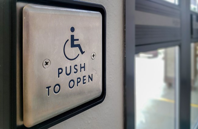 Square metal button with wheelchair logo, with glass door beyond.