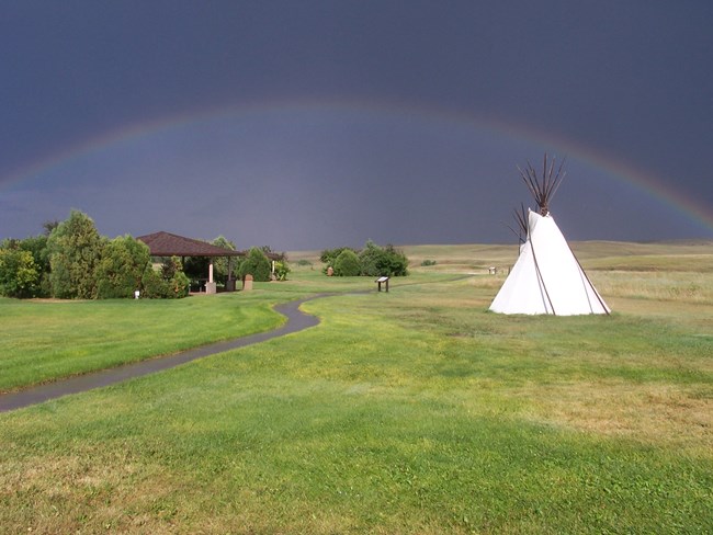 A rainbow arcs over prairie, a wet walkway, pavilion, and white tipi against a dark sky.