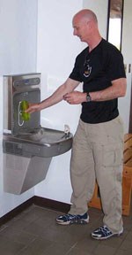 A man in black shirt and tan pants holds a small water bottle under a metal water dispenser/fountain.