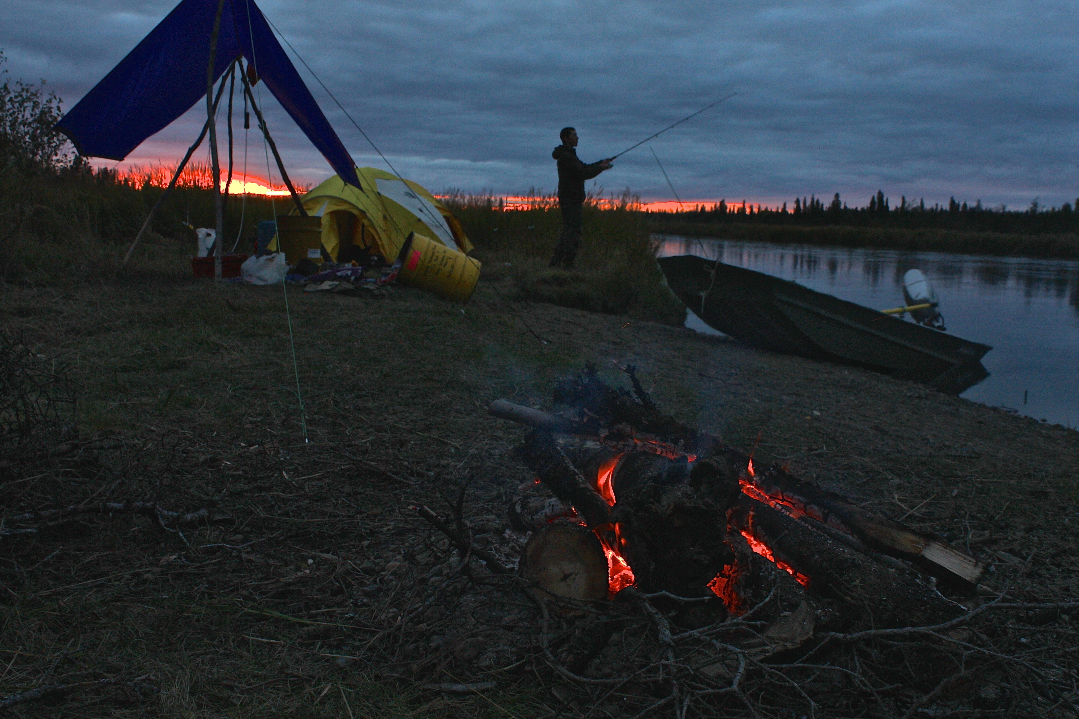A man at a campsite fishing with fire, boat and tent in scene