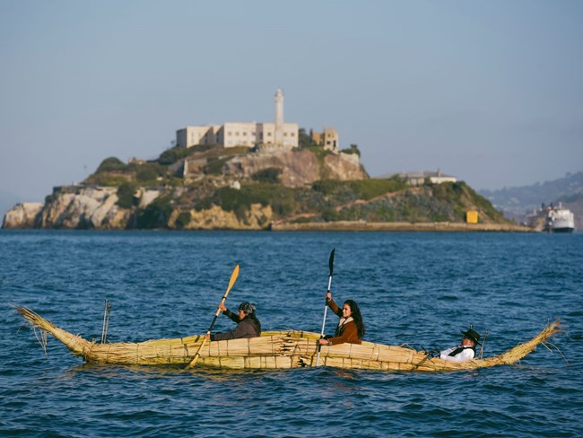 Three people paddle in canoe made of dried tule reeds, Alcatraz Island in the background.