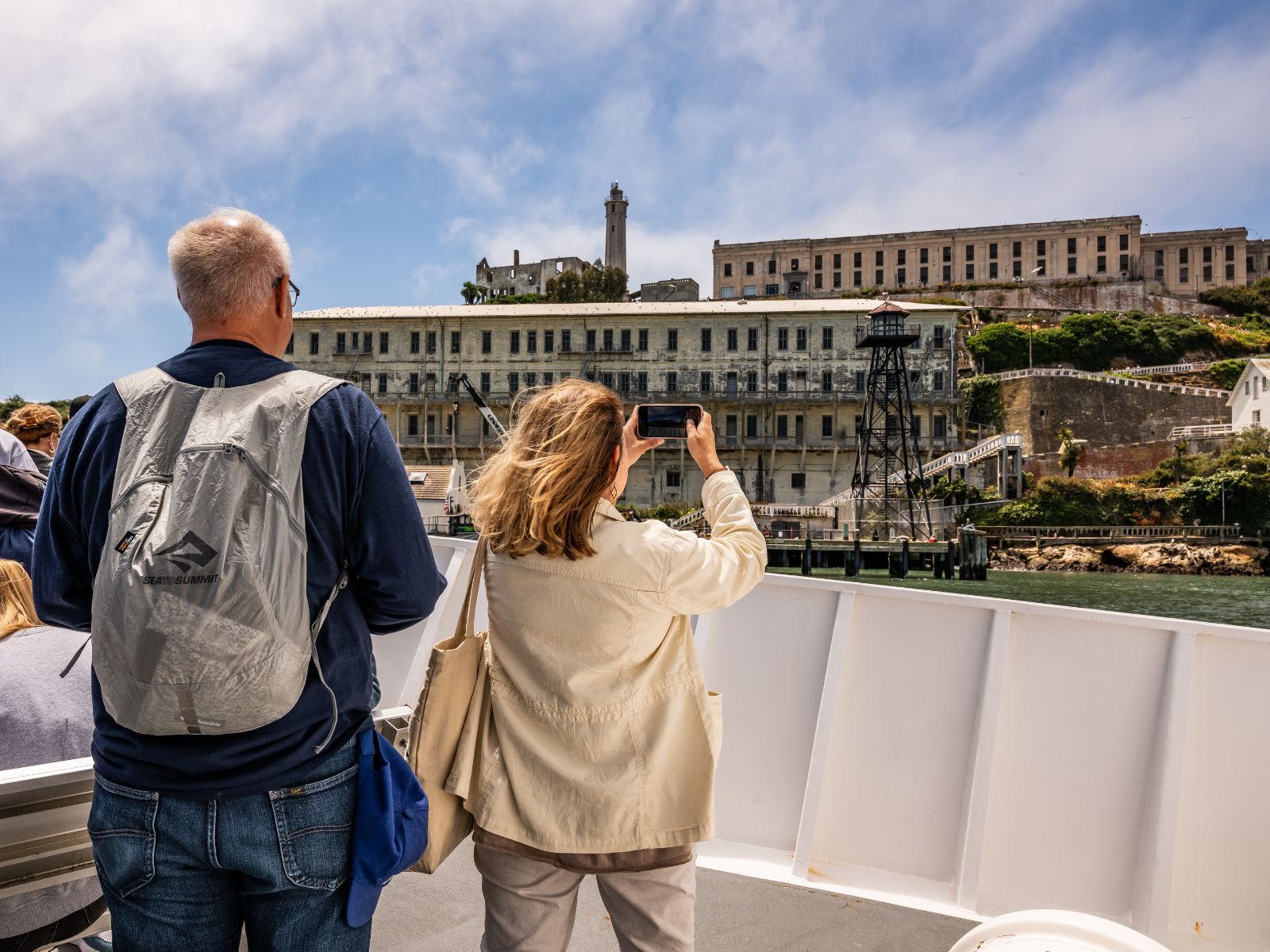 Two visitors on the bow of a boat, taking pictures of a large building on the Alcatraz dock.