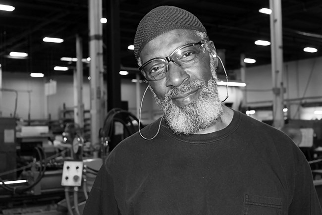 Ameen, Retired Aerospace Tech, wears clear safety glasses and crochet cap in what looks like a factory/warehouse. Included with permission from Pioneer Human Services.