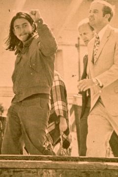 Indigenous man being escorted of Alcatraz by white men in suits, raising fist in air.