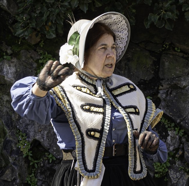 A woman wearing a historical bonnet, shirt, and skirt gestures with her hands.