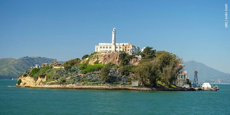 Lighthouse sits atop tree covered island. Dock, guard tower along at left shore.