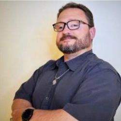 Bearden man in black framed glasses and denim button-down shirt smiles at camera, arms crossed in front of chest.