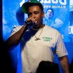 John Cannon Black man in white t-shirt and green baseball cap speaks into a microphone in front of a crowd.