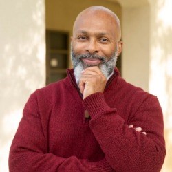 Ernst Fenelon Smiling Black man with salt and pepper beard, bald head, and red sweater looks directly at camera.