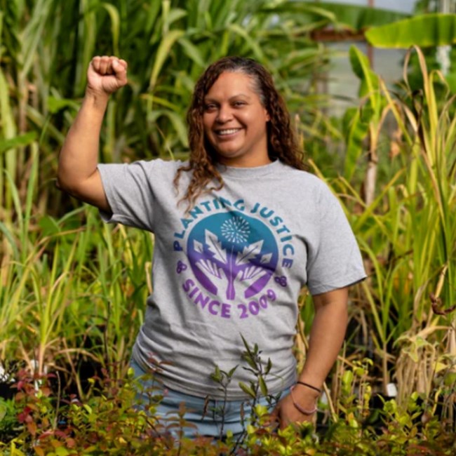 Curly-haired woman stands among plants, right fist raised. Shirt reads “Planting Justice since 2009”