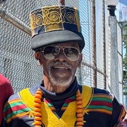 Black man in yellow shirt with goatee smiles at camera. He wears a black cap and glasses.