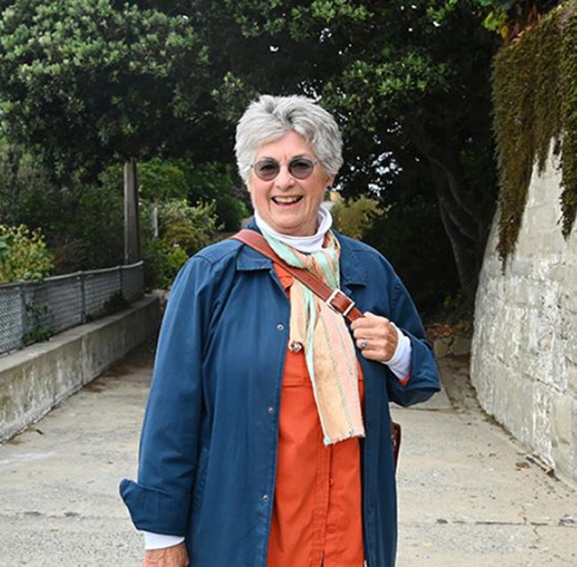 A woman with gray hair, sunglasses, and blue and orange shirts smiles on a road surrounded by greenery.