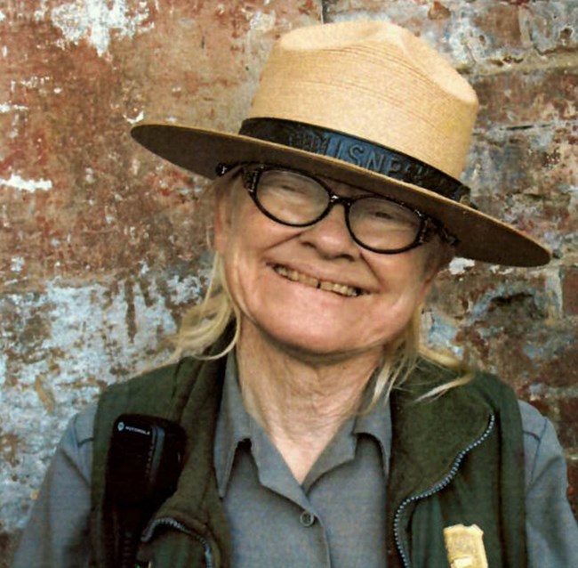 A woman in a ranger uniform, flat hat, and glasses smiles at the camera.