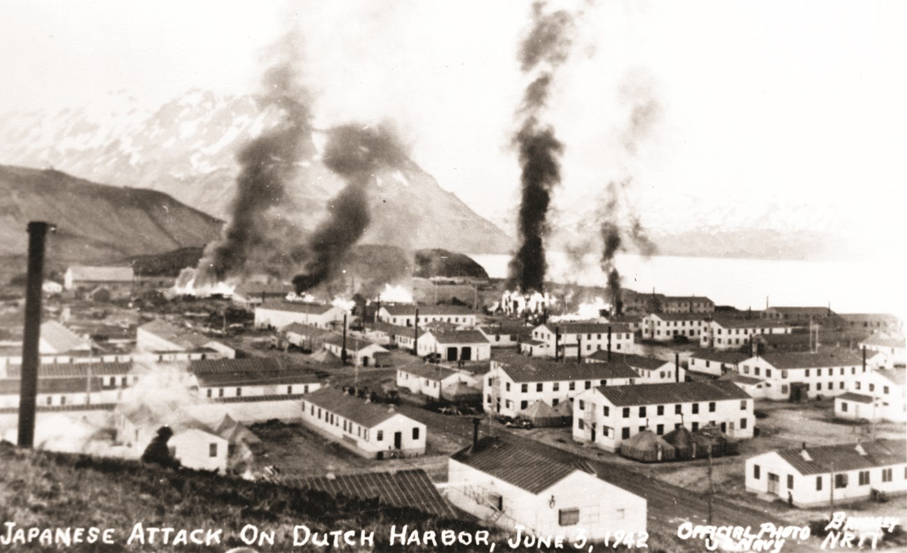 Many white two-story white buildings in a town, with four columns of black smoking rising from four locations.