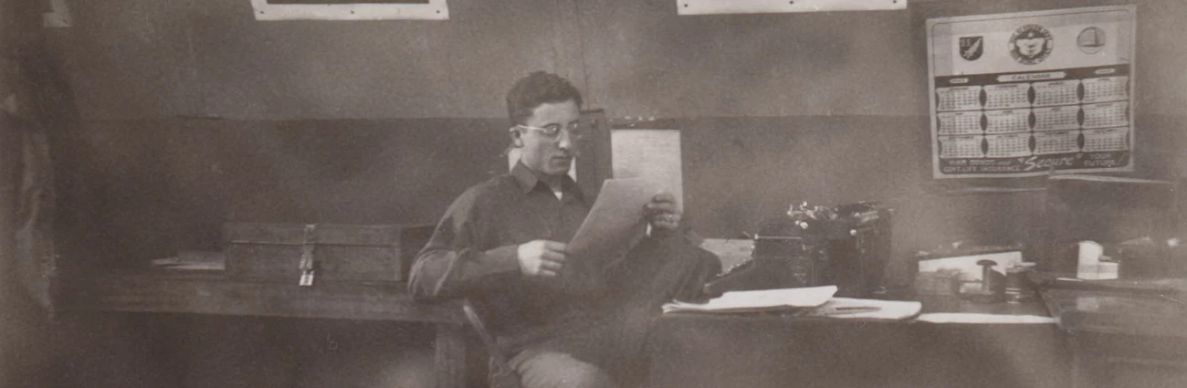almartinreading in a historic scene, a young man with glasses reads a paper sitting at a messy desk with war posters on the wall behind him.