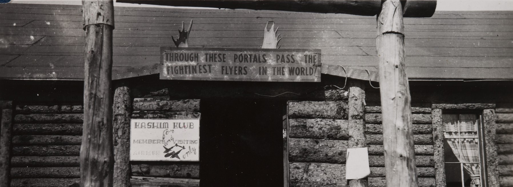 a historic scene of an open door with two items hanging above the frame: a pair of moose antlers and a sign reading "through the portals pass the fightin-est flyers in the world."