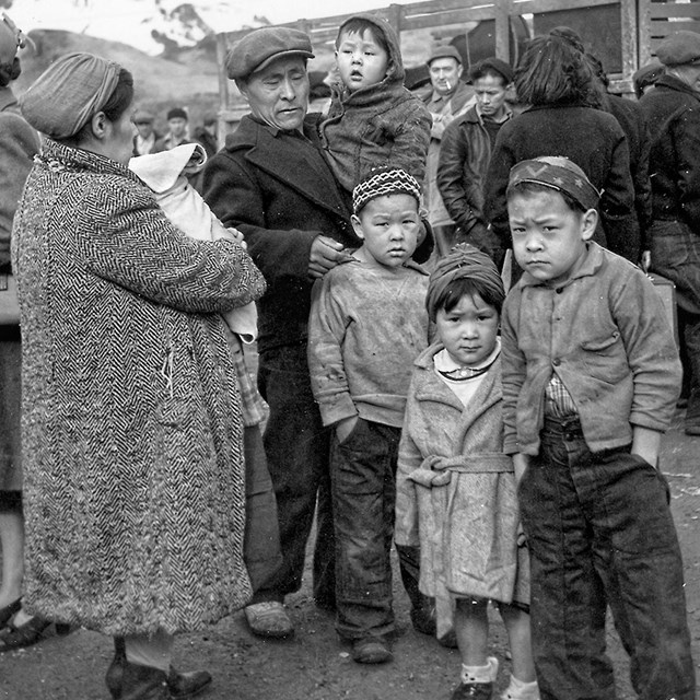 A historic scene of a family of seven standing amid a large crowd of other people, all wearing hats and warm clothing.