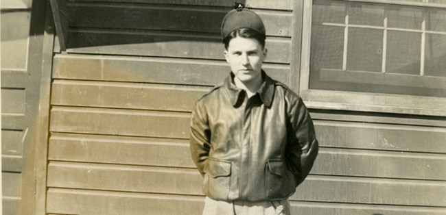 lt= A black and white photo of a man facing the camera in a bomber jacket and hat.
