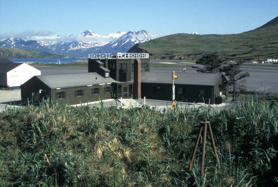 A dark colored building with brick obelisk out front and mountains in the background.