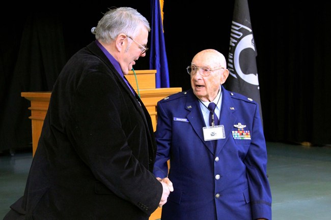 A man in a suit coat shakes hands with a man in a blue uniform