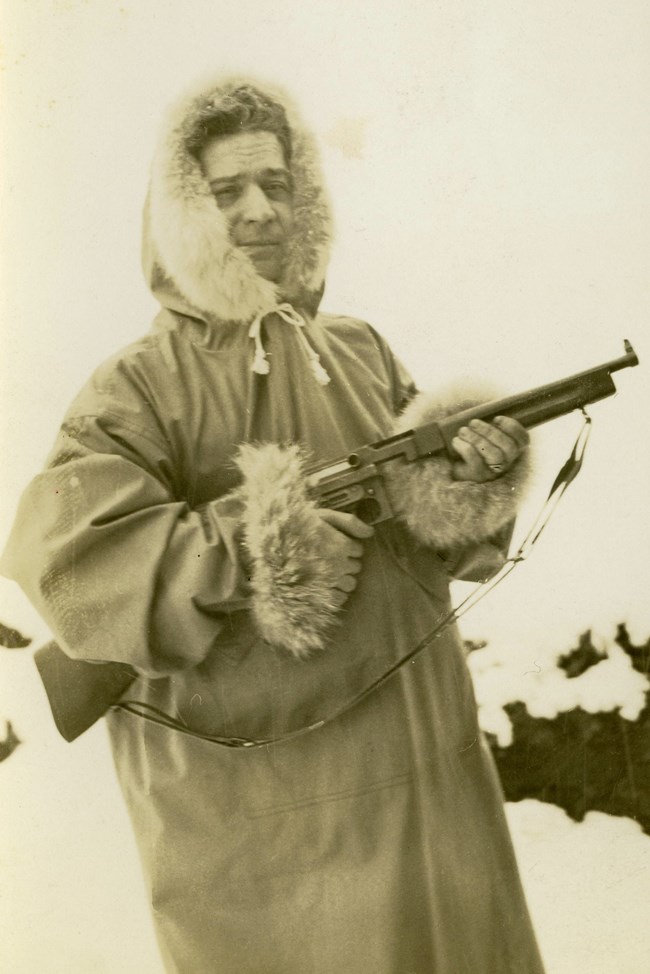 in a historic scene, a man holding a gun wears a long, thick winter coat with a fur-lined hood and cuffs.
