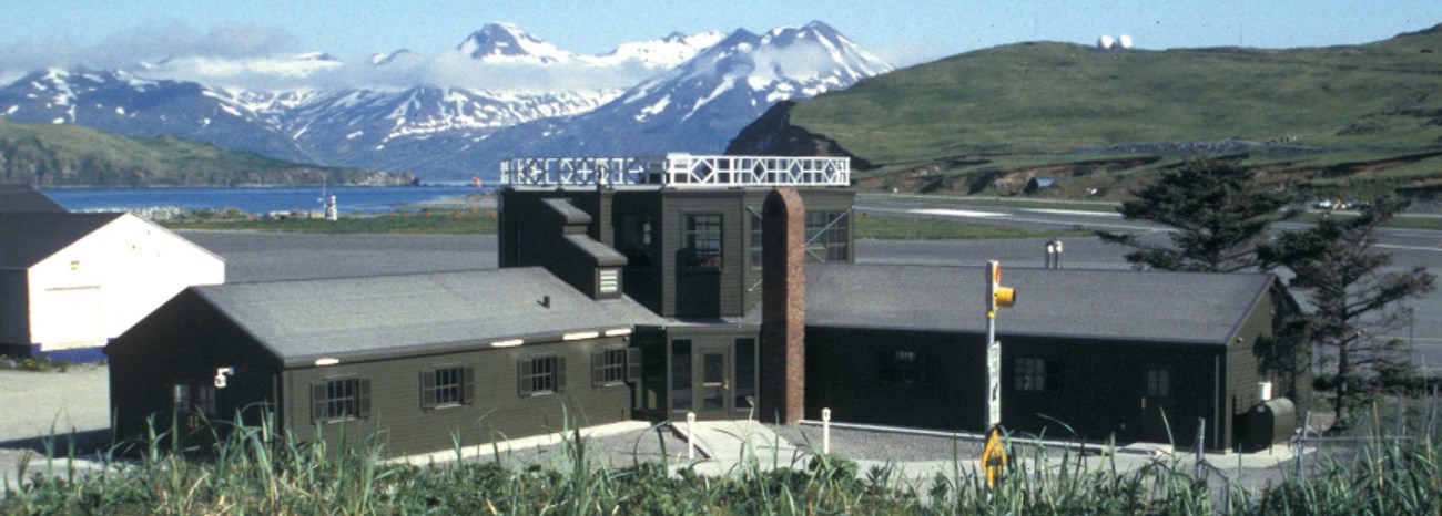 an L-shaped building with a tall, central lookout sits in front of a coastal mountain range.