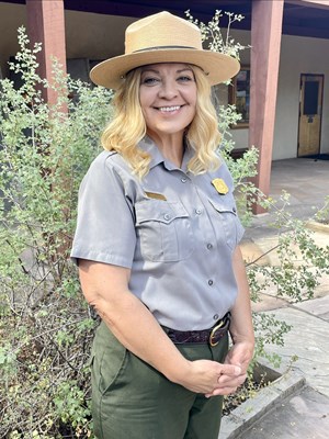 A female park ranger stands in front of a building with some shrubbery.