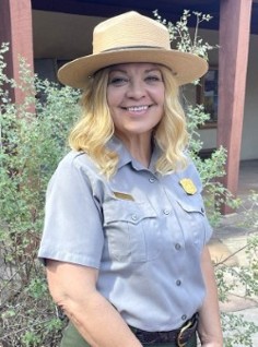 Smiling female park ranger wearing a tan wide-brimmed hat and gray uniform standing outdoors near greenery.