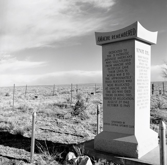 Black and white image of "Amache Remembered" monument located within the Amache cemetery. Monument is in the foreground and is surrounded by fence posts and is among the sagebrush and other high plains desert vegetation.
