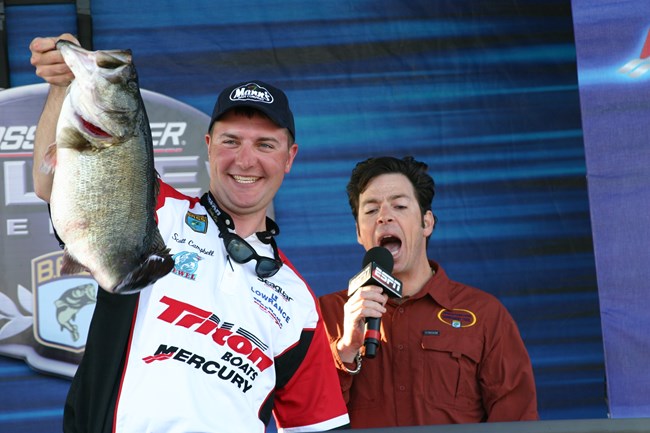 Image from the 2007 Bassmaster Elite Series at Lake Amistad with man holding bass by lower lip and announcer in background.
