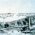 Black and white image of a bridge being built high above a river canyon with a segment still missing near the center and construction equipment and materials visible above and below.