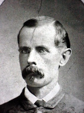 Headshot photo of a man in his late-20s with bushy mustache, high brow, piercing gaze, and civilian clothes.