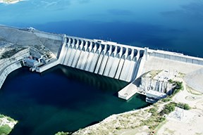 Amistad Dam with what appears to be a full reservoir of deep blue water behind it. There are hydroelectric generator buildings on either side with a pool of water in front between the three structures.