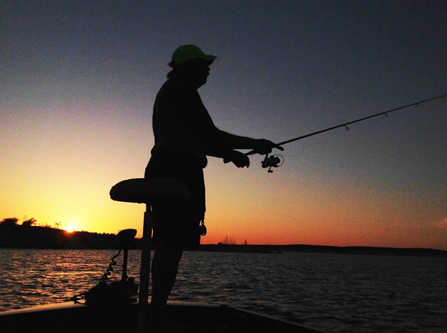 Silhouette of person fishing from boat at sunrise.