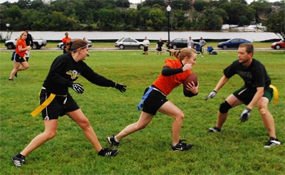 A woman runs with a football and is pursued by two defenders in a flag football game on a grassy field.