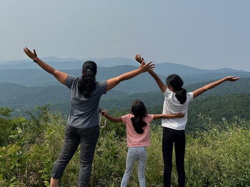Three women, the middle one shorter, standing facing away from the camera. Their arms are raised as if they are embracing the beautiful view of the hills and forestry they are facing.