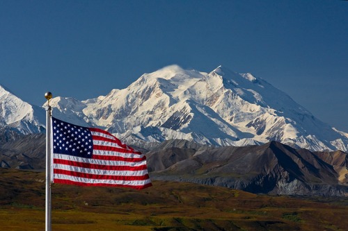 The American Flage waving in the bottom left corner in front of Mt. Mckinley against a clear blue sky.