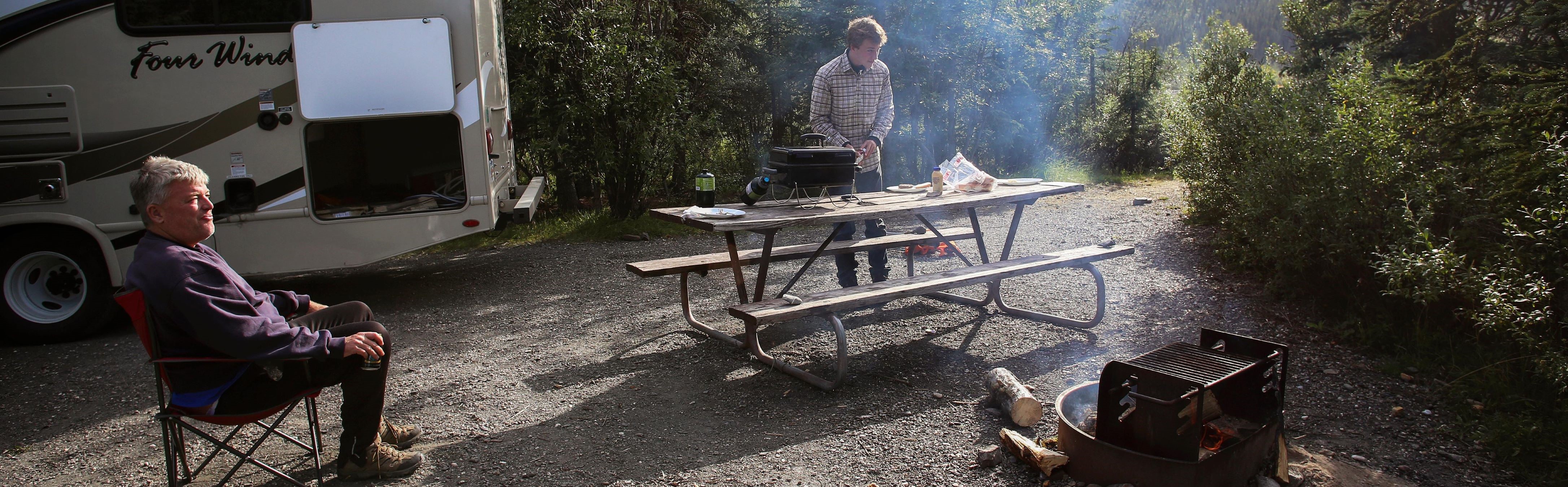 Campsite; back of RV, person in camp chair, person at picnic table, campfire.