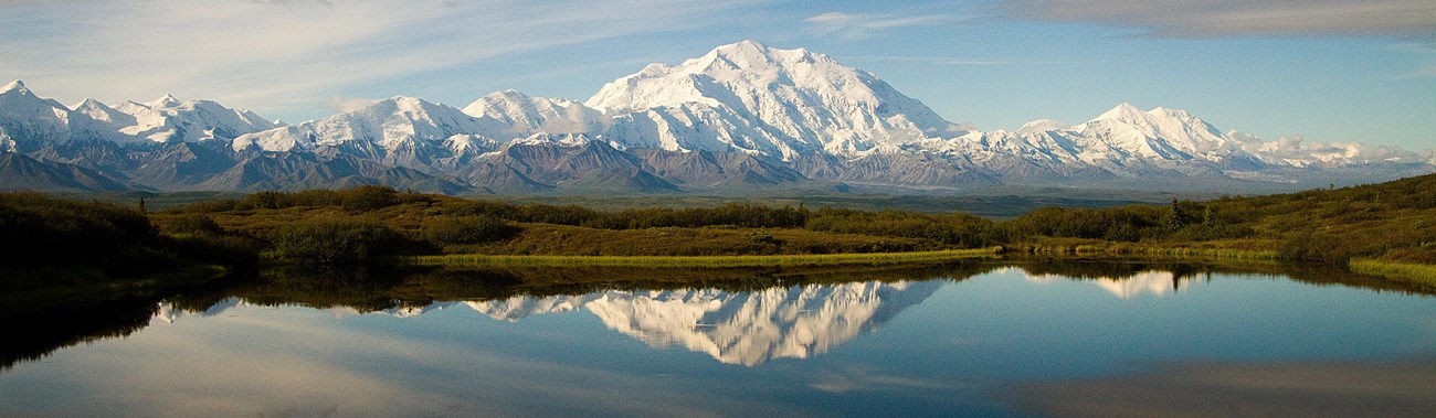 Large snow covered mountain in center of photo with lake reflecting it in foreground.