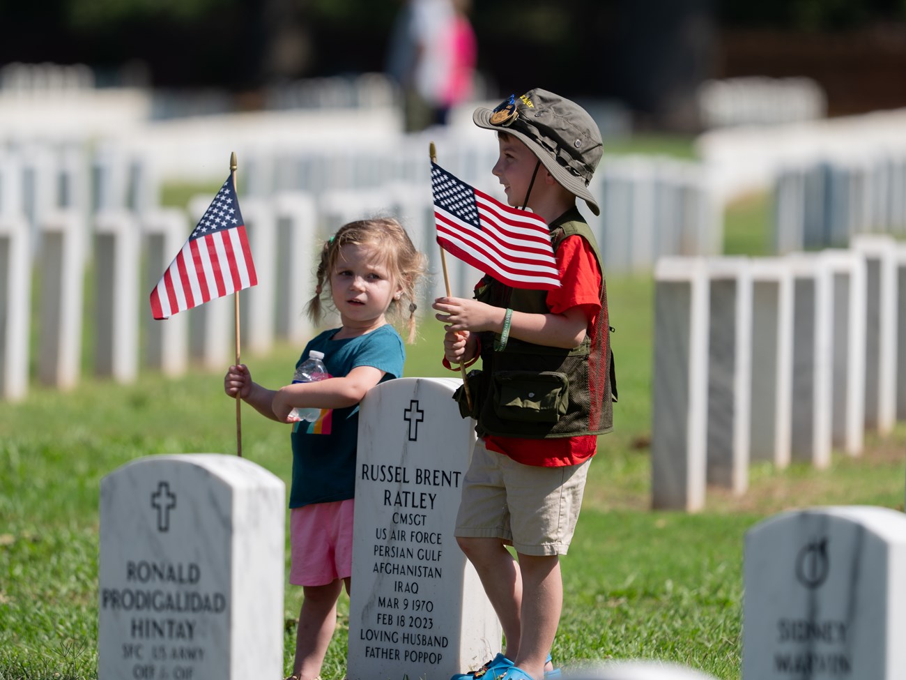 Two kids holding American flags standing amongst headstones decorated with American flags.