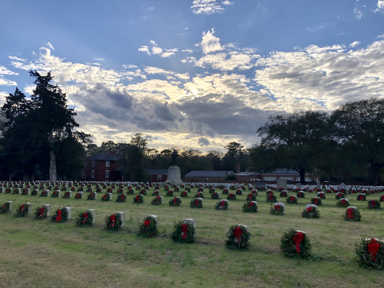 thumbnail_IMG_9120 Sunset photo with Christmas wreaths on headstones in a cemetery. Large brick building in the background
