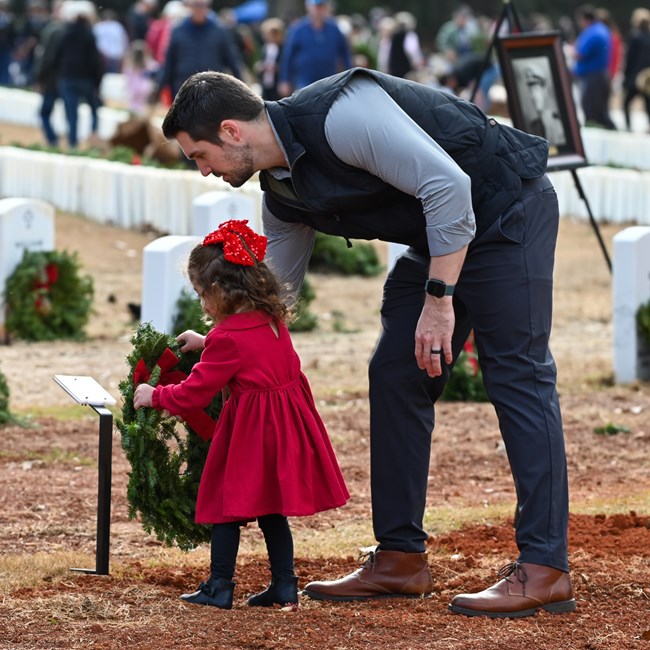 A father and daughter place a decorative wreath in front of a headstone.