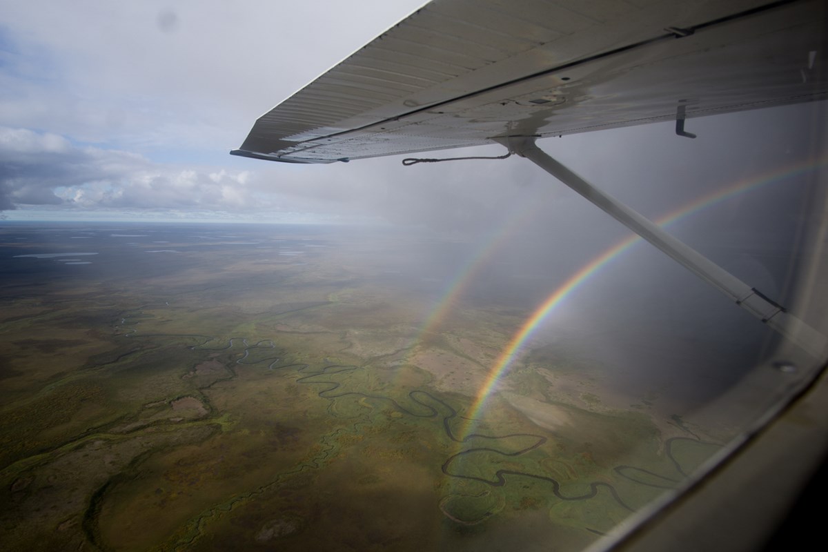 a view from a small aircraft over vast tundra with a meandering river; a double rainbow arcs beyond the wing of the plane