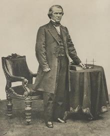 Andrew Johnson standing by a table and chair with his hand on a book