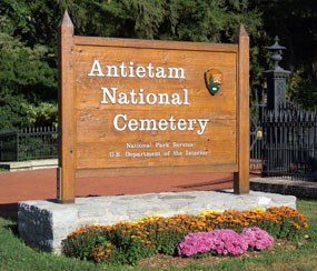 A rectangular, wooden sign reads "Antietam National Cemetery" with a National Park Service logo in the corner.