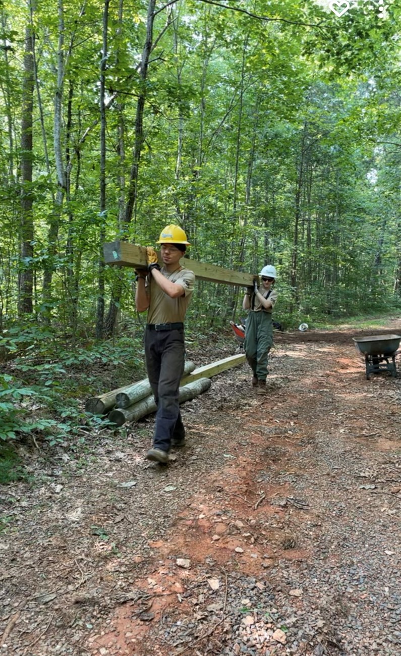 Two youth trail crew members carry a long wooden beam across their shoulders on a trail with trees in the background.