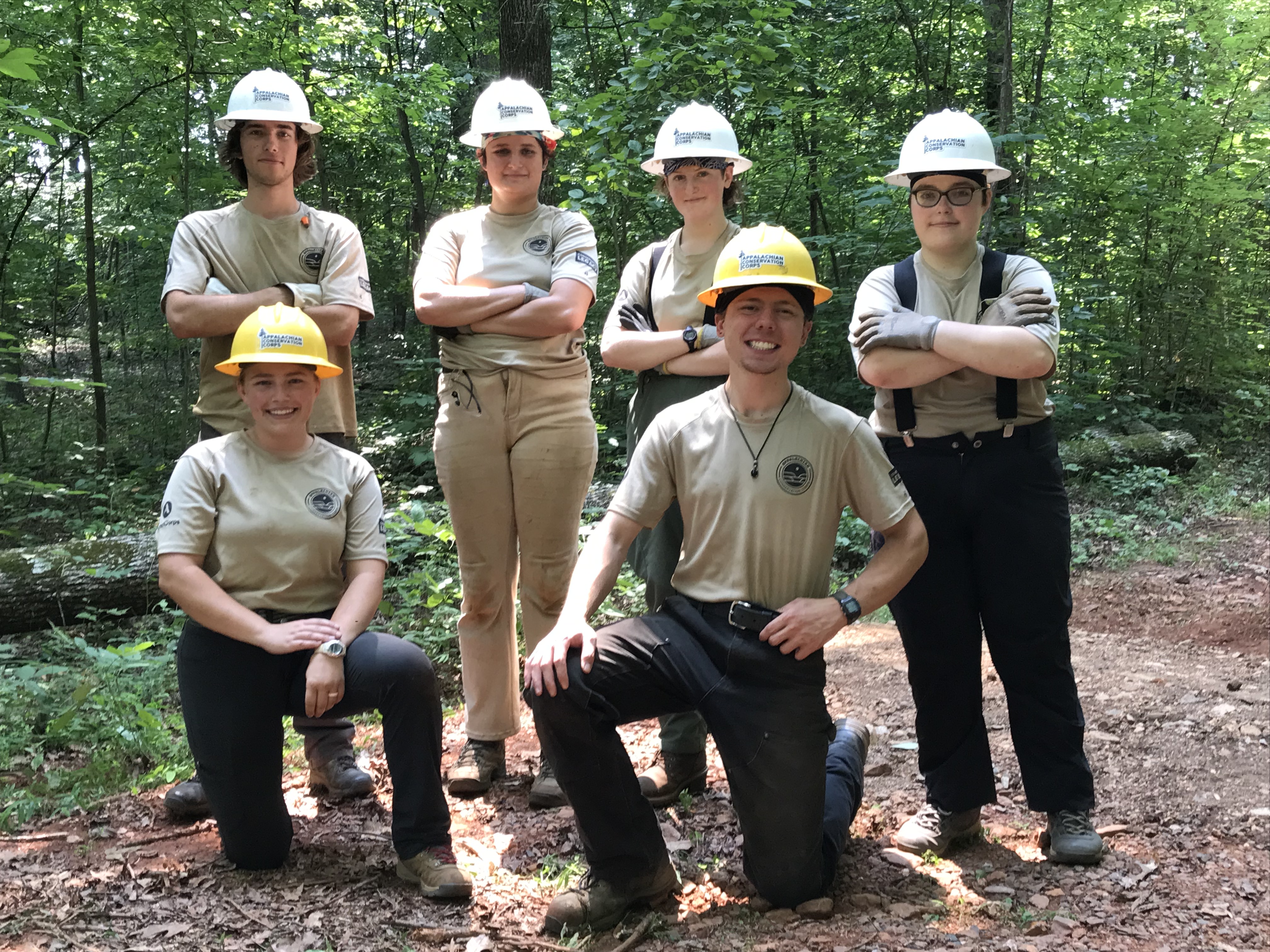 Six young trail crew members wearing khaki and hard hats stand as a group with arms folded
